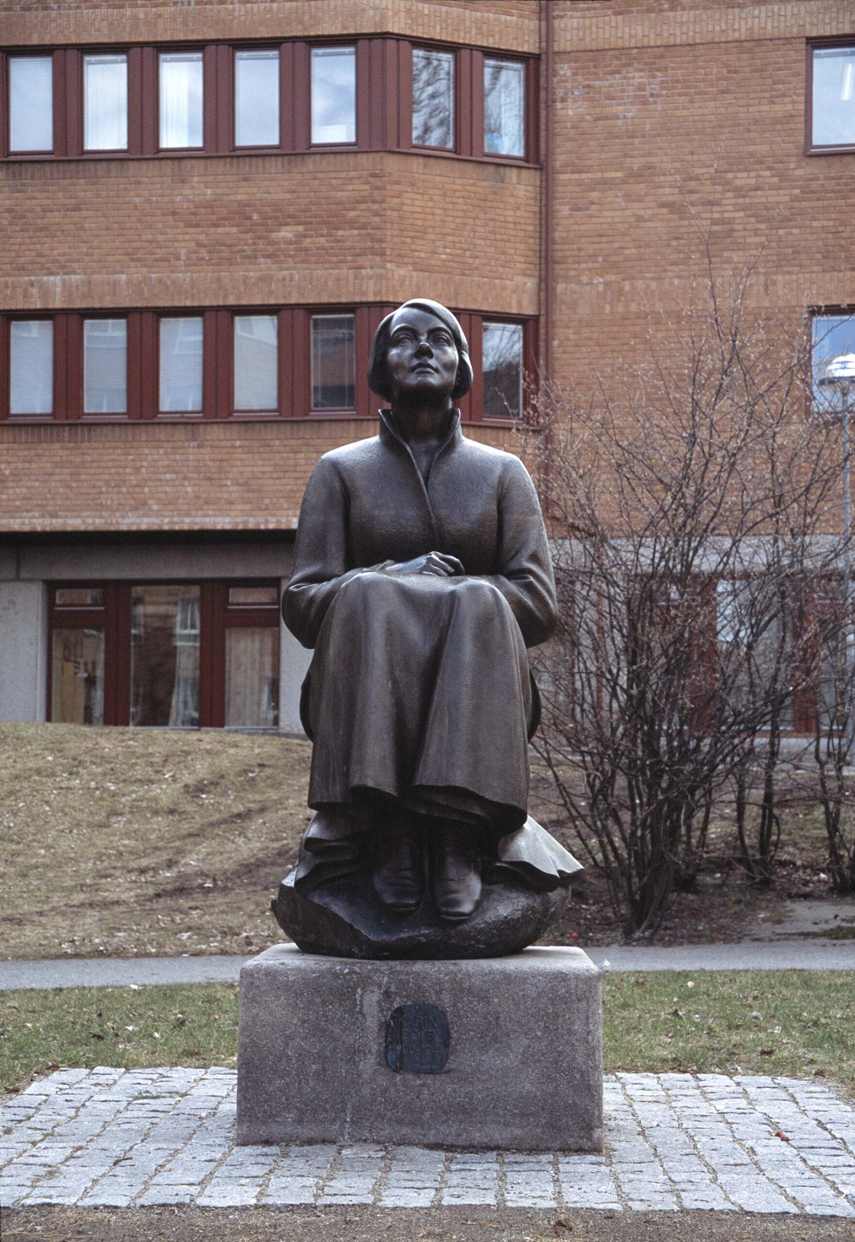 En skulptur av en kvinna som sitter på en sten. Utomhus med tegelhus bakom.