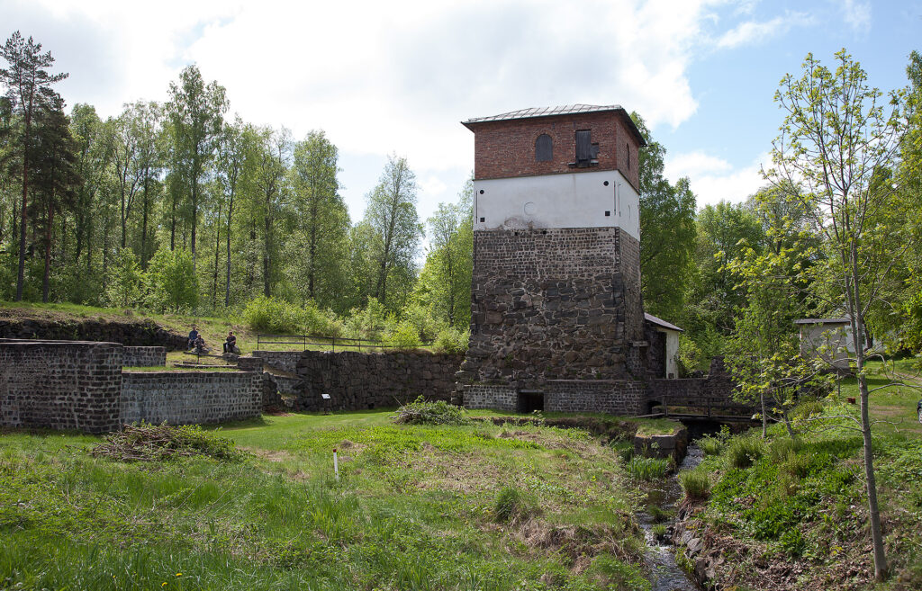 En fyrkantig byggnad står vid en liten bäck. Det är sommar.