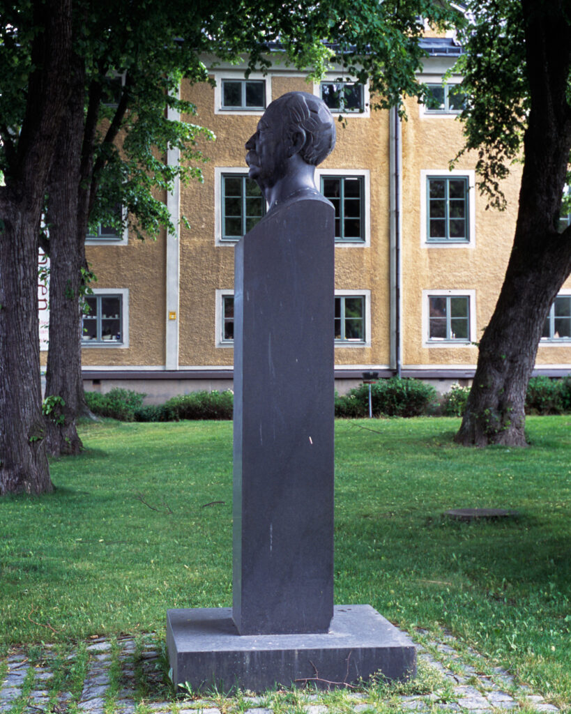 Ett ansikte i profil av sten på en pelare. Gräs runtom och ett gult hus i bakgrunden.