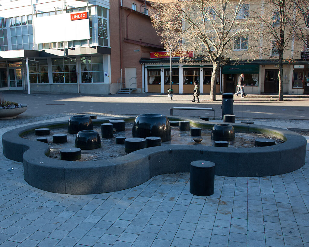 En fontän med svarta kanter och bubblig form på ett torg. Hus bakom.