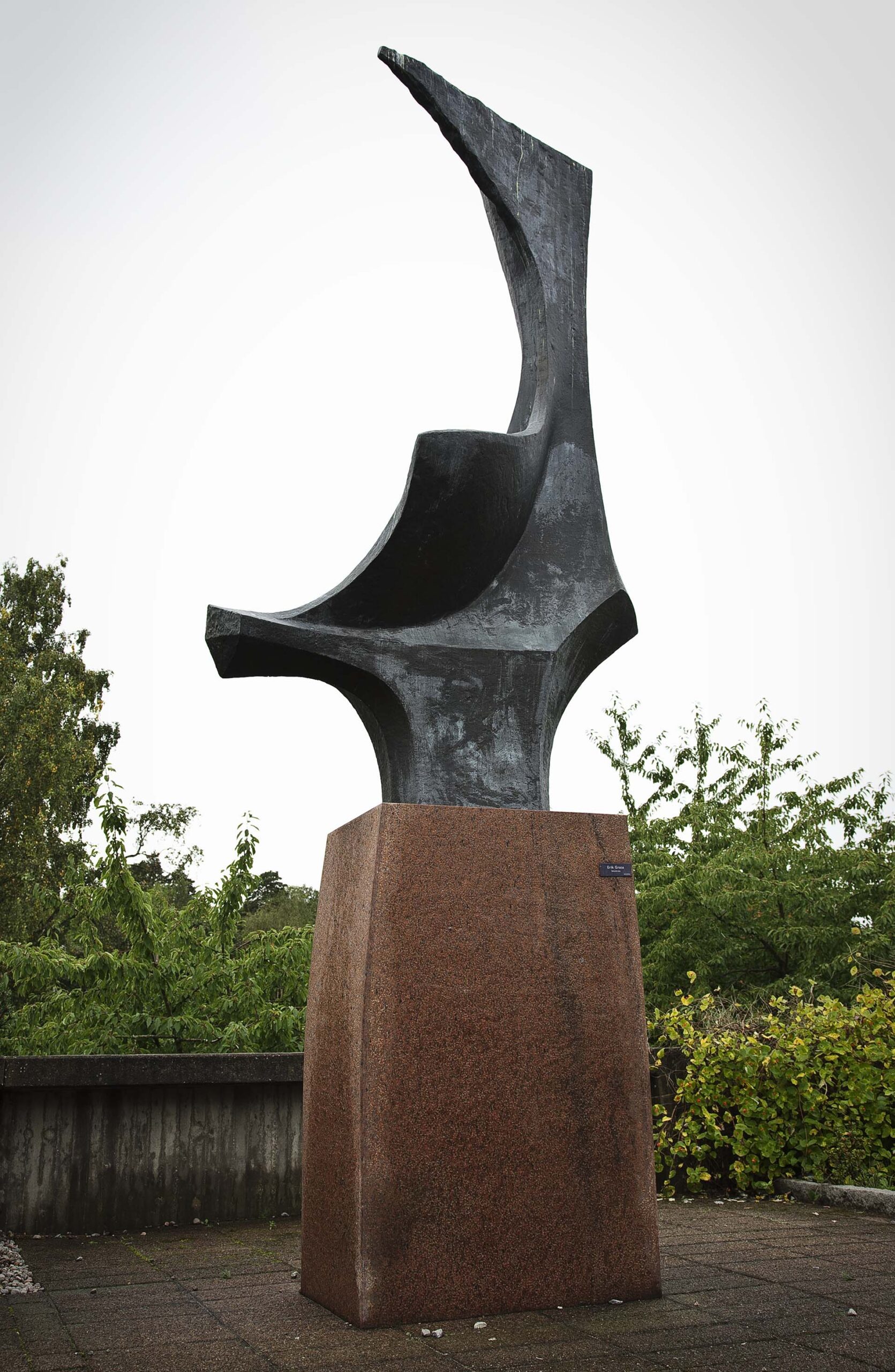 En skulptur i brons med abstrakt form på en hög sockel av röd sten. Himmel och grönska bakom.