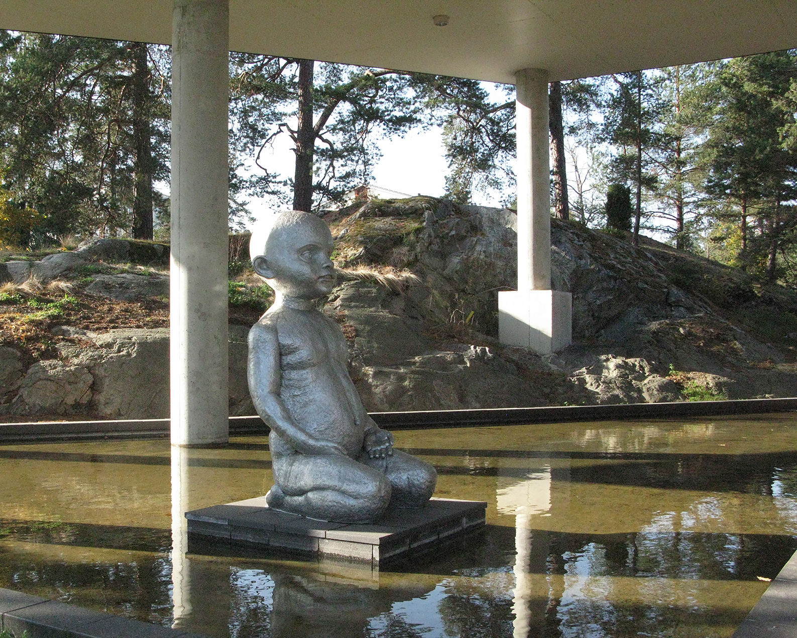 En skulptur av en flicka gjord av sten sitter i en fontän. Träd bakom.