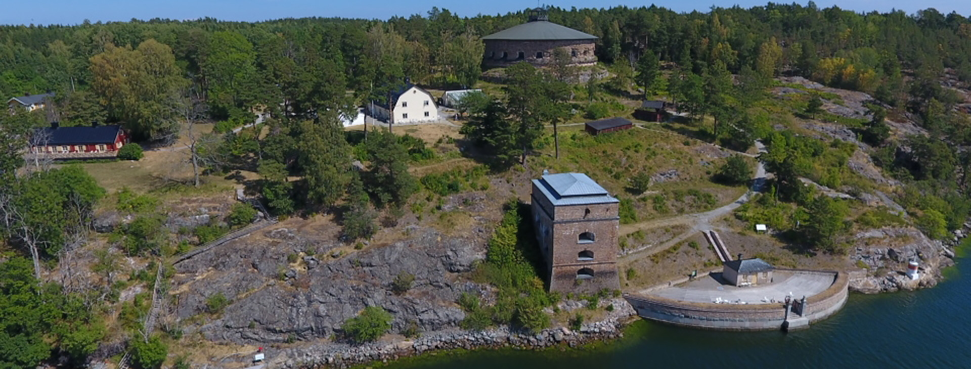 Vy över hus som står på en ö, det är buskar och hav.