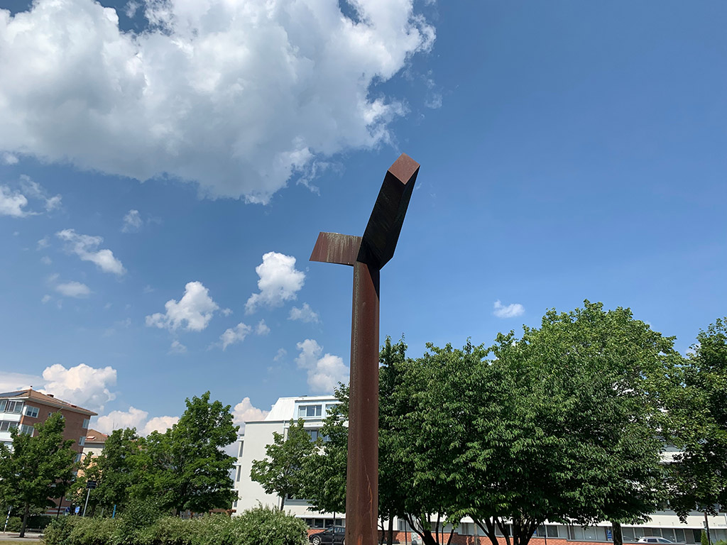 Glänsande skulptur i rostigt stål framför tegelbyggnad.