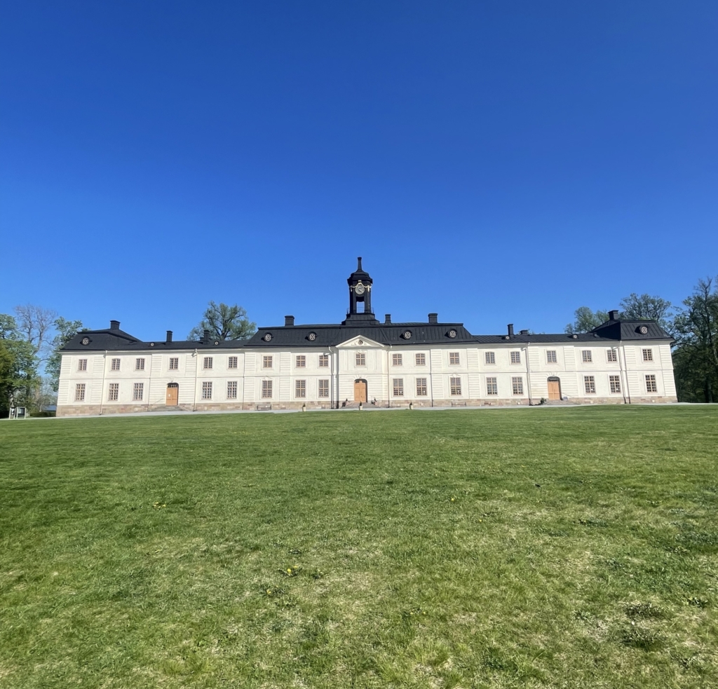 Foto av det slottet som brer ut sig i sin fulla längd framför en grön gräsmatta med en klarblå himmel som fond.