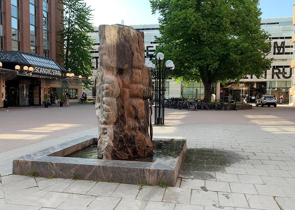 Abstrakt stenskulptur med vatten i en bassäng på ett torg.