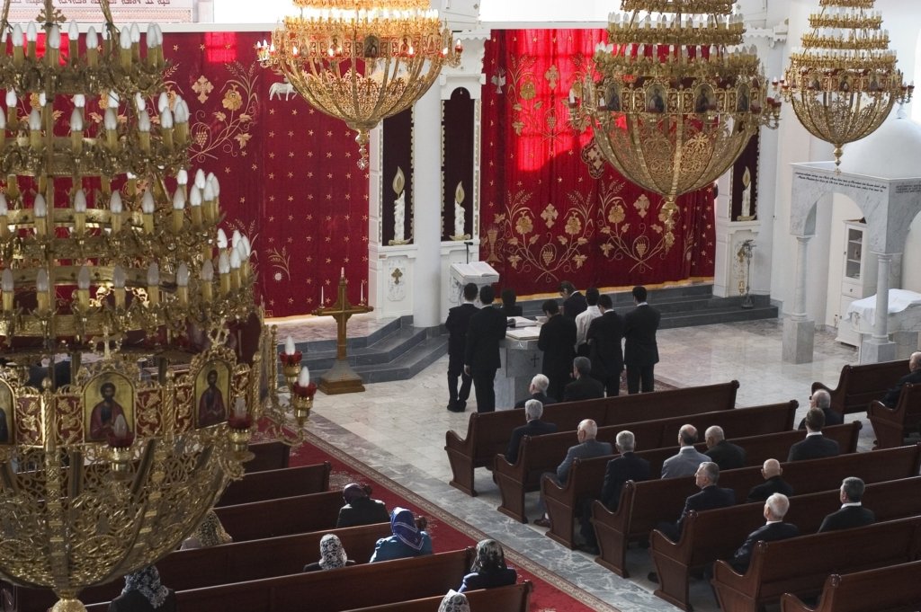 Foto taget från läktaren visar röda draperier hänger längst fram i kyrkorummet, stora takkronor av mässing och människor som sitter i bänkraderna.