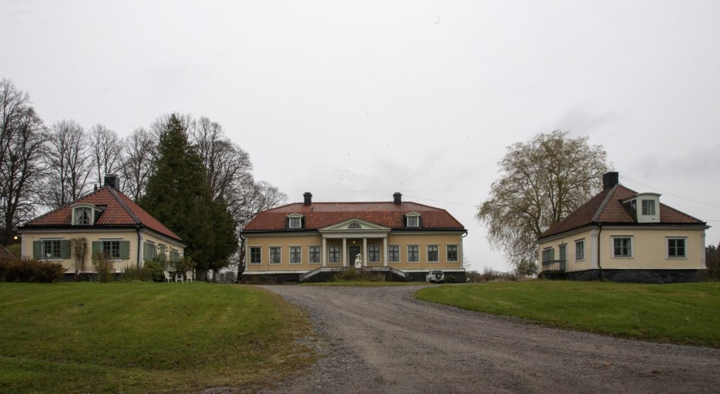 En grusväg leder fram till den gula huvudbyggnaden med brutet tak. Den flankeras av två flyglar.