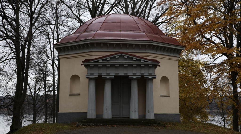 Templet i ljusgul kulört har ett välvt vinrött koppartak och ligger på en kulle. med vattnet bakom. Entrén markeras av fyra grå pelare.