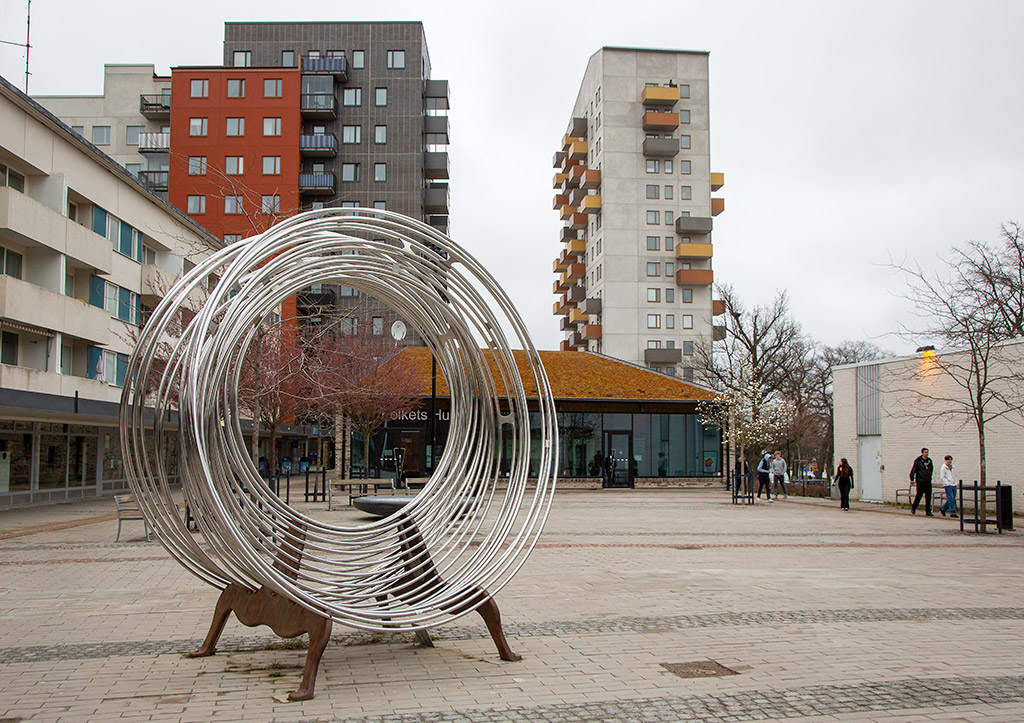 Skulptur med ringar av rostfritt stål med ben av järn som står på ett torg.