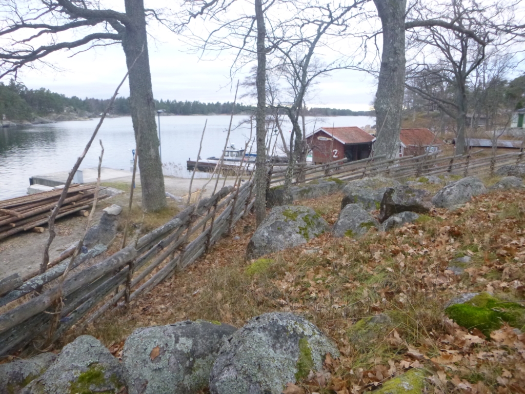 Stenbumlingar och en gärdsgård bland höstlöv i förgrunden och några röda hus, vattnet och en båt i bakgrunden