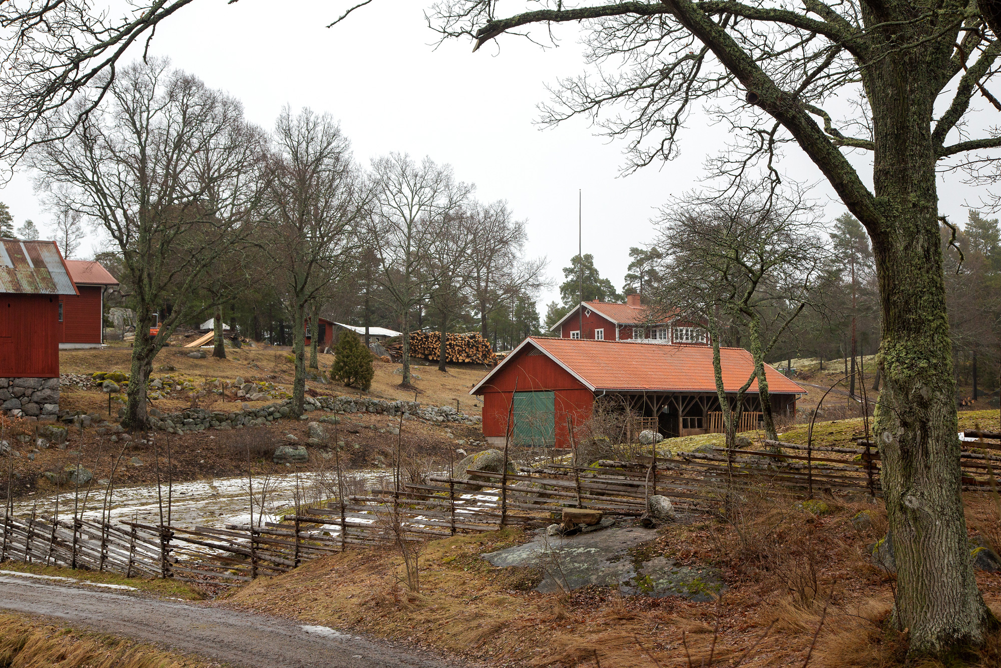 I förgrunden syns en gärdsgård och bakom den ett landskap med flera rödmålade byggnader.