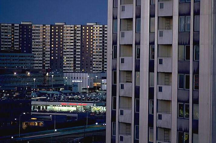 En skymningsbild där ett höghus i förgrunden tar upp halva bilden. Bakom syns centrumet med flera lampor och Storstugan, det stora bostadshuset som en skärm bakom.