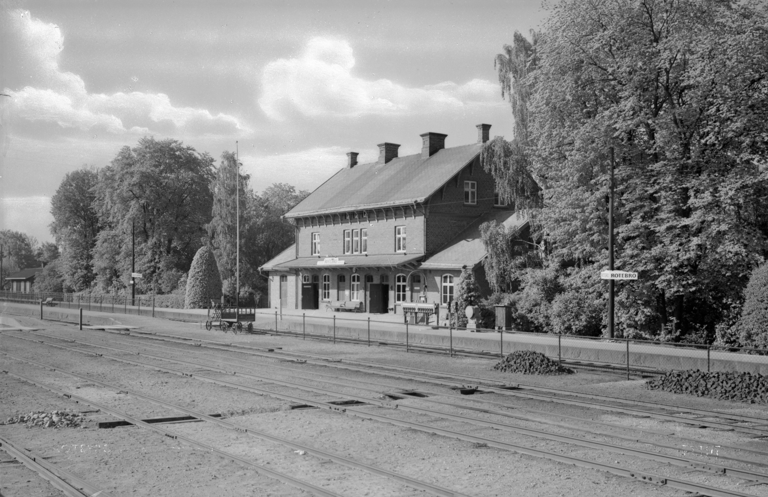 Ett svartvitt foto av stationshuset med perrong och spårområde.